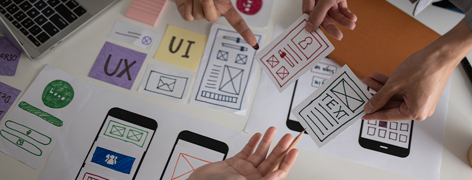 Team collaborating on mobile app design with UI and UX wireframes, sketches, and layout prototypes spread on a desk.